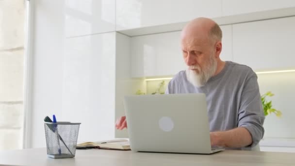 Lindo hombre de 70 años de edad, de pelo gris con barba a la edad de usar el ordenador de navegación por Internet, la lectura de un libro electrónico en la aplicación se comunica en línea a través de enlace de vídeo con el médico. Educación a distancia hombre viejo. — Vídeo de stock