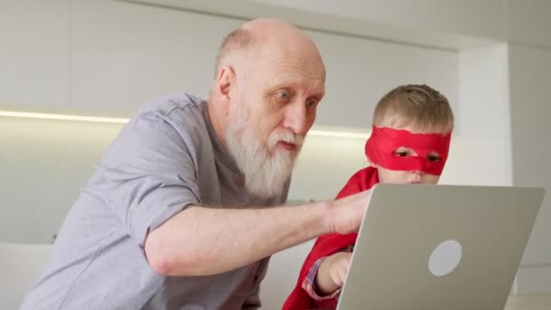 Äldre farfar med grått skägg, tillsammans med pojke sonson i röd regnrock, sitta vid datorn och titta på tecknade serier, kommunicera via videolänk med släktingar, chatta och läsa nyheter på laptop hemma. — Stockvideo