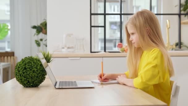金髪の女の子はノートパソコンで家で宿題をしている。オンライン学習、遠隔授業、自宅での教育、学童のための技術、ビジネス会議。子供たちは家で宿題をする。学校に戻る. — ストック動画