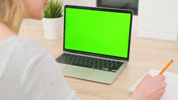 Estudiante trabaja con computadora portátil de pantalla verde cromakey, utilizando el teclado para escribir texto y bolígrafo para escribir información en el bloc de notas en la sala de estar de la casa. — Vídeos de Stock