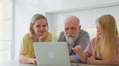 Gülümseyen genç bir kadın ve kızı yaşlı bir babaya evde dinlenirken laptopta uygulamaları nasıl kullanacağını gösteriyor. 70 yaşında, kır saçlı, yaşlı bir emekli modern dizüstü bilgisayar kullanmayı öğreniyor..