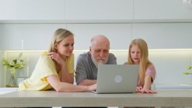 70 yaşında yaşlı, kır saçlı, yaşlı bir emekli modern dizüstü bilgisayar kullanmayı öğreniyor. Gülümseyen genç bir kadın ve kızı yaşlı bir babaya evde dinlenirken laptopta uygulamaları nasıl kullanacağını gösteriyor..
