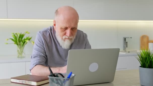 Lindo anciano viejo trabaja en casa detrás de la computadora portátil. Abuelo jubilado aprende a usar el teclado portátil y posar mientras está sentado en casa en el lugar de trabajo. Nueva tecnología para personas mayores. — Vídeo de stock