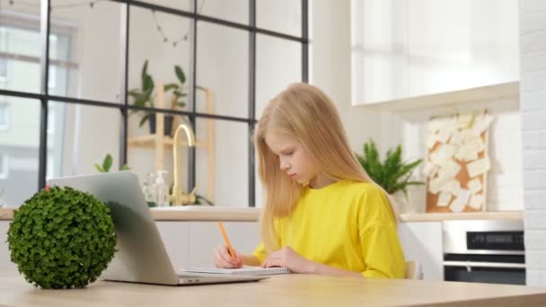 Blond školačka se učí online pomocí notebooku, dělat si poznámky do notebooku. Teenage školačka se dívá na internetový video kurz, zatímco sedí u svého domácího stolu. Koncepce distančního vzdělávání. — Stock video