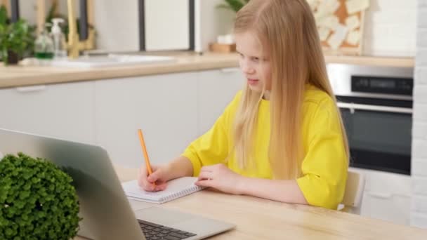 ブロンドの女子高生はノートパソコンを使ってオンラインで学習し、ノートブックにメモを取る。十代の女子高生は自宅のテーブルに座りながらインターネットビデオコースを見ています。距離学習の概念. — ストック動画