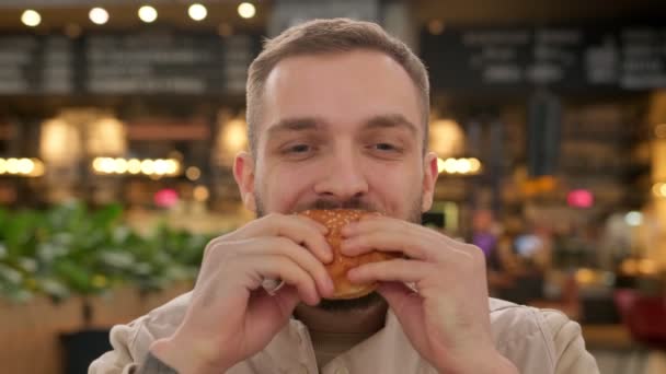 Unshaven caucasian man eats meat burger with pleasure, bites burger with his mouth, man casually eats junk food from fast food, smears his face with ketchup sauce on background of fast food restaurant — Vídeo de Stock