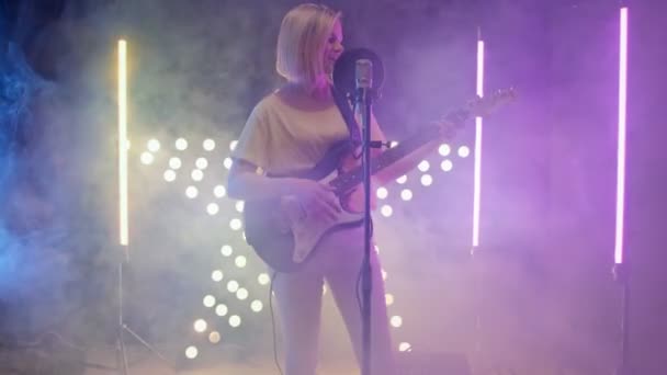 Blond girl artist singer sings song into microphone and plays an electric guitar filled with light and smoke room. Musicians records song in recording studio. Caucasian artist performs. — Stock Video