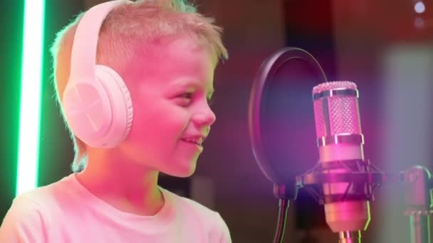 Biały dzieciak nagrywa nowy utwór muzyczny, śpiewa piosenkę do mikrofonu. Proces nagrywania piosenki w studiu nagraniowym. Portret młodego blond piosenkarza noszącego słuchawki, śpiewającego piosenkę w studiu nagraniowym. — Wideo stockowe