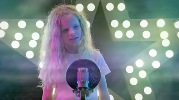 Young girl singer sings karaoke song standing on stage against background of glowing multicolored lights. Cheerful smiling girl on stage of karaoke club sings her favorite songs in rays of club lights — Stock Video