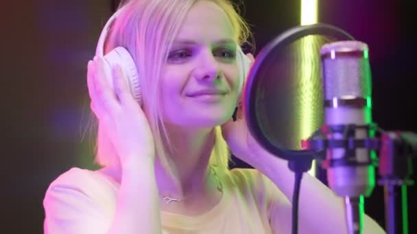 Jeune fille blonde artiste star chanteuse enregistre chanson à succès en studio d'enregistrement, va au microphone met des écouteurs, danse et chante chanson dans le microphone dans la chambre noire avec des lumières multicolores scintillantes. — Video