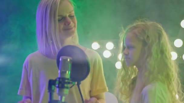 Ξανθά κορίτσια παίζουν τραγούδια καραόκε σε πάρτι στη σκηνή σε κλαμπ. Χαρούμενη μητέρα και κόρη τραγουδούν σε νυχτερινό κέντρο διασκέδασης με πολύχρωμα φώτα. — Αρχείο Βίντεο