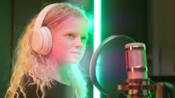 Młoda piękna dziewczyna zdejmuje słuchawki z głowy i powiesza je na stojaku z mikrofonem i pozostawia ramkę. Dziewczyna nagrywa swój głos w studiu nagraniowym. — Wideo stockowe