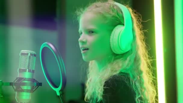 Młoda blondynka śpiewa do mikrofonu w białych słuchawkach w profesjonalnym studiu nagraniowym w kolorowych promieniach światła. Uśmiechnięty piosenkarz śpiewa piosenki do mikrofonu w ciemnym studio. — Wideo stockowe
