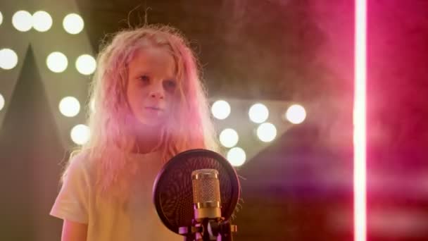 Cheerful smiling girl on stage of karaoke club sings her favorite songs in rays of club lights. Young girl singer sings karaoke song standing on stage against background of glowing multicolored lights — Stock Video