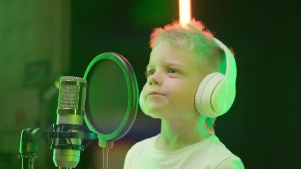 Portret młodego blond piosenkarza noszącego słuchawki, śpiewającego piosenkę w studio nagraniowym. Biały dzieciak nagrywa nowy utwór muzyczny, śpiewa piosenkę do mikrofonu. Proces nagrywania piosenki w studiu nagraniowym. — Wideo stockowe