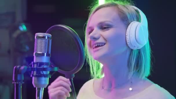 Une fille caucasienne enregistre une nouvelle piste musicale, chante une chanson dans un microphone. Portrait fille blonde chanteuse dans un casque chantant chanson dans un studio d'enregistrement de musique. — Video