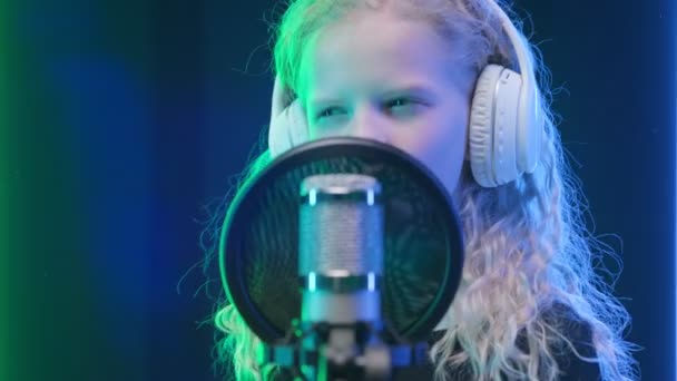 Joven chica caucásica graba nueva pista de música, canta canción en el micrófono. Retrato chica rubia cantante en auriculares cantando canción en estudio de grabación de música. — Vídeo de stock