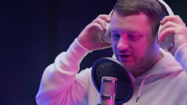 Rappeur caucasien non rasé portant un casque en sweat-shirt blanc ou un sweat à capuche, enregistrant une chanson hip-hop devant le microphone dans un studio d'enregistrement professionnel rempli de lumière au néon colorée. — Video