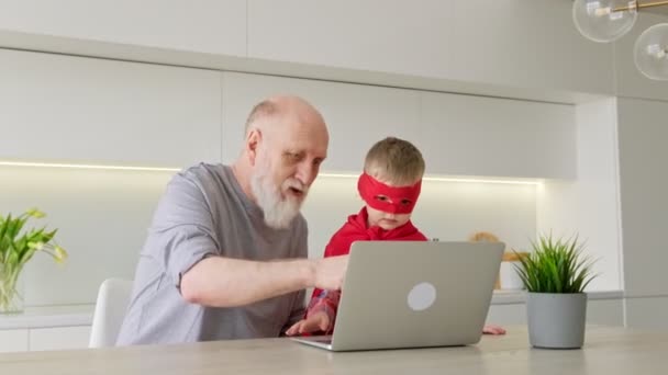 Pequeño niño rubio junto con un abuelo de edad avanzada pasar el tiempo libre, comunicarse y jugar a la computadora portátil moderna, ver dibujos animados sentados mesa en casa. El ocio familiar del abuelo y el nieto. — Vídeos de Stock