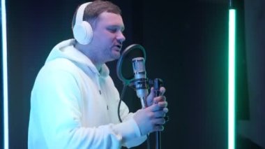 Beyaz sweatshirt ya da kapüşonlu kulaklık takan, profesyonel kayıt stüdyosunda, renkli neon ışıklarla dolu bir mikrofon önünde hip-hop şarkısı kaydeden, traşsız, beyaz tenli rapçi..