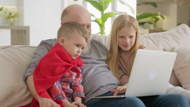 Szczęśliwe pokolenie starszy siwowłosy dziadek ze swoimi wnukami, wnuczką i wnukiem siedzą na kanapie na laptopie i komunikują się ze swoimi krewnymi z czatu wideo i dobrze się bawią. — Wideo stockowe