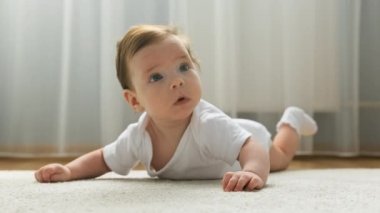Sevimli bir kız bebek yerde yüzüstü yatıyor, kameraya bakıyor ve gülümsüyor. Erken bebek gelişimi. Bebek odasında yerde yatıyor, halının üzerinde sürünüyor..