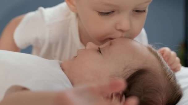 Заботливый братец нежно и с любовью целует свою двухмесячную сестру, лежащую на кровати на лбу. Концепция отношений в счастливой семье между братом и сестрой, брат помогает заботиться о младшей сестре. — стоковое видео