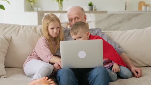 Šťastná generace starý dědeček a vnoučata, chlapec a dívka, sedět společně u notebooku na gauči doma, komunikovat prostřednictvím video spojení s příbuznými. Děti učí starší osoby práci na notebooku — Stock video