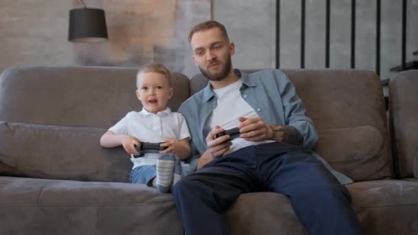 Happy family bearded father and fair-haired young son play video games on console, hold joysticks, press buttons, look at camera, sitting at home. Happy family and concept of modern technologies. — Vídeo de stock