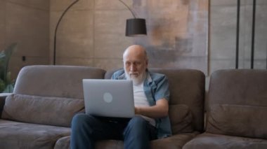 Emotions of winning in pensioner, winning in casino, winning in sports betting, winning in video game. Old grandpa, happy emotionally raising his hands up, looking at laptop screen, sitting on couch.