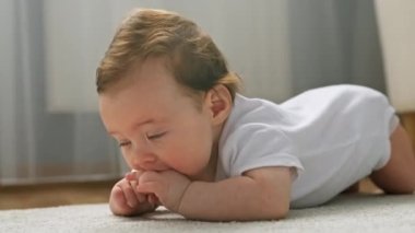 Portrait of cute newborn baby in white bodysuit, lying on his stomach on living room carpet, sucking his fingers, looking interestingly at camera with his mouth open and his legs dangling.