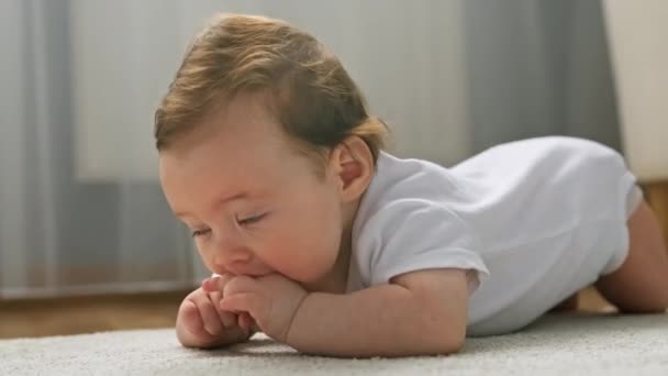 Portrait of cute newborn baby in white bodysuit, lying on his stomach on living room carpet, sucking his fingers, looking interestingly at camera with his mouth open and his legs dangling. — Stok video