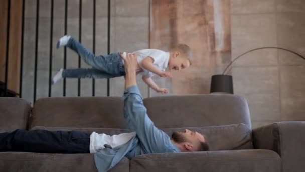 Lindo chico rubio juega juego con su padre en avión, vuela en brazos de su padre, niño vuela como avión extiende sus brazos a un lado. Feliz relación familiar entre padre e hijo. — Vídeos de Stock