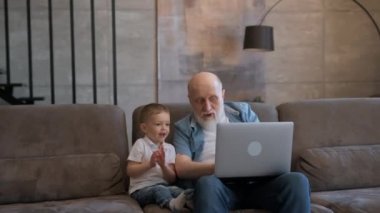 Mutlu yaşlı büyükbaba ve tatlı küçük torun gülerek ellerini kaldırarak dizüstü bilgisayara bakıyorlar, mutlu zaferler, evdeki kanepede oturuyorlar. Büyükbaba ve çocuk oyunu kazanmaktan mutluluk duyar..