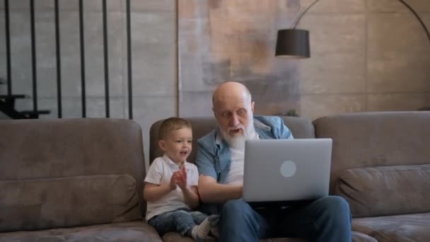 Velho avô feliz e pequeno neto bonito rindo, feliz levantando as mãos olhando para o laptop, vitória feliz, sentado no sofá em casa. Avô e filho estão felizes em ganhar ou ganhar jogo. — Vídeo de Stock