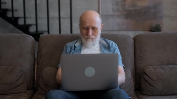 Yaşlı kişi internette dizüstü bilgisayarda çalışıyor, haberlere bakıyor, internet tarayıcısında arama yapıyor, uzaktan evde çalışıyor. Yaşlı iş adamı büyükbaba bilgisayarda çalışıyor. Evdeki koltukta oturuyor.. — Stok video