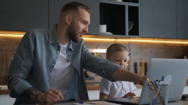 Šťastná rodina, mladý vousatý otec spolu se světlovlasým synem, pomocí notebooku studovat a surfovat v internetovém prohlížeči. Otec pomáhá svému malému synovi s domácím úkolem, zatímco sedí u kuchyňského stolu.. — Stock video