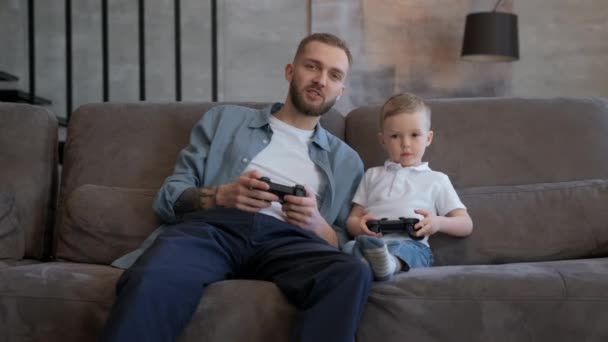 Retrato de pai e criança jogando videogame no sofá em casa, pressionando botões no joystick falando olhando para a câmera. Família feliz e conceito de tecnologia moderna. — Vídeo de Stock
