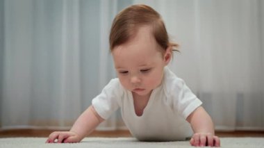 Erken bebek gelişimi. Bebek odasında yerde yatıyor, evde halıda sürünüyor. Sevimli bir bebek yerde yüzüstü yatıyor, kameraya bakıyor ve gülümsüyor..