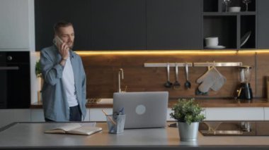 Genç adam ev ofisinden uzaktan çalışıyor mutfakta yürüyor ve müşterilerle ya da evdeki müşterilerle iletişim kurarken telefonla konuşuyor. Adam ev ofisinden üstleriyle telefonda konuşuyor..