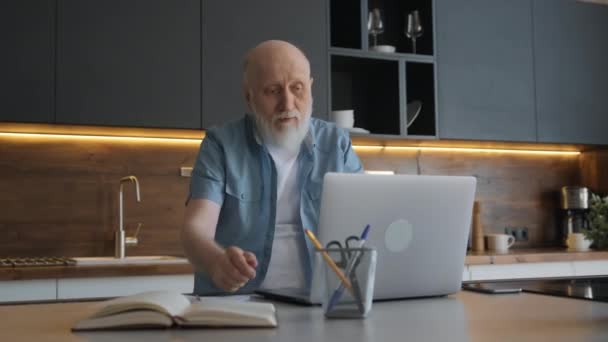 Yakışıklı yaşlı adam, dizüstü bilgisayarda çalışıyor ve görüntülü sohbet zoom 'u ile konuşuyor, merkezdeki masada oturuyor. Happy Man modern dizüstü bilgisayar kullanıyor. Aile ve arkadaşlarla çevrimiçi iletişim. — Stok video