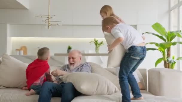 Avô idoso com seus netos se divertir, se divertir com seus netos, filhos saltar e abraçar o avô, se divertir em família em casa. Crianças saltam no sofá abraçando avô aposentado. — Vídeo de Stock