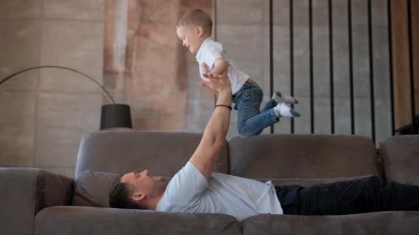 Cámara lenta feliz familia papá levanta niño para volar como avión, divertirse, disfrutar de fin de semana mañana soleada, acostado en el sofá, despreocupado adorable pequeño bebé jugando con el padre-padre en casa. — Vídeo de stock