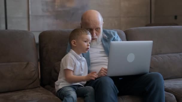 Літній літній чоловік разом зі своїм онуком сидить разом на дивані з ноутбуків, махаючи вітаннями руками, співрозмовником по відеозв'язку. Спілкування з сім'єю за допомогою відео чату . — стокове відео