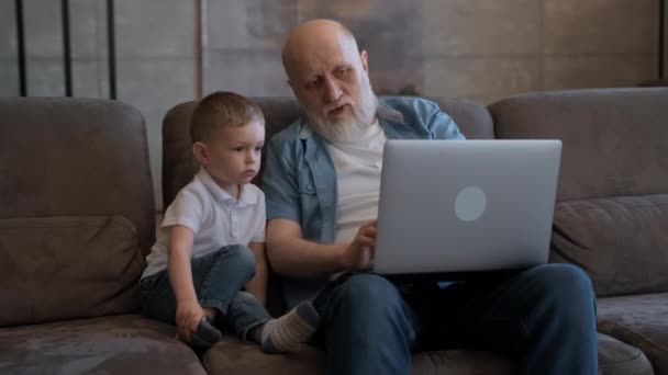 Дід старого спілкується з молодим онуком, сидячи на ноутбуці, дивлячись мультфільми та відеочати збільшуючи, сидячи на дивані, сімейне з'єднання двох поколінь за допомогою ноутбука . — стокове відео