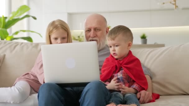 Семья поколений, внуки и пожилой старик на пенсии смотрят на экран ноутбука, смотрят видео, общаются через видео-чат через веб-камеру, сидят на диване в светлой комнате. Счастливого семейного времяпровождения. — стоковое видео