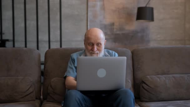लॅपटॉपवर उत्साही वृद्ध आजोबा दिसतात, इंटरनेटवर यश साजरे करतात, घरी सोफ्यावर बसतात. अनुफोरिक मनुष्याला नवीन दूरस्थ नोकरी मिळते, चांगली बातमी ईमेल वाचून विजयाचा आनंद होतो, प्रेरणा मिळते . — स्टॉक व्हिडिओ