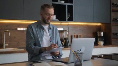 Tıraşsız erkek serbest çalışıyor. Evde dizüstü bilgisayarının başında oturuyor. Web kamerasıyla görüntülü sohbet yoluyla iletişim kuruyor. Öğrenci çevrimiçi bir öğretmenden öğrenir, konuşur ve not defterine metin yazar. 