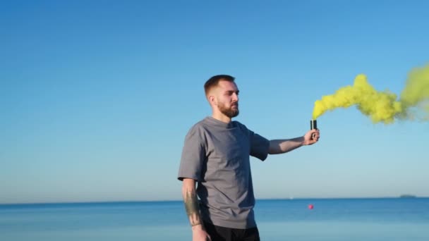 수염을 기른 젊은 남자 가 해변을 따라 푸른 하늘 과 바다를 배경으로 노란색 연기와 유색인 폭탄을 들고 걷고 있습니다. 사람은 해변에서 연막탄 과 함께 노란 연기 신호 폭탄을 발사 한다. — 비디오