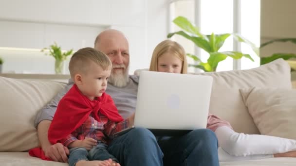 Nesiller boyu aile, torunlar ve yaşlı bir adam dizüstü bilgisayara bakıyor, video izliyor, web kamerasıyla iletişim kuruyor, parlak odadaki kanepede oturuyor. Mutlu aile eğlencesi. — Stok video